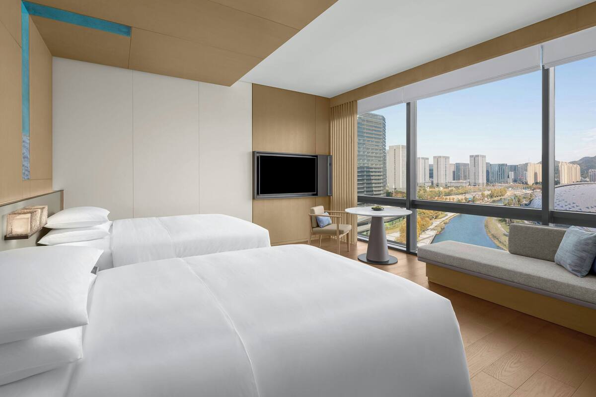 Ein Hotelzimmer mit zwei Betten, einem Fernseher, einem kleinen Tisch und einem großen Fenster mit Blick auf die Stadt.