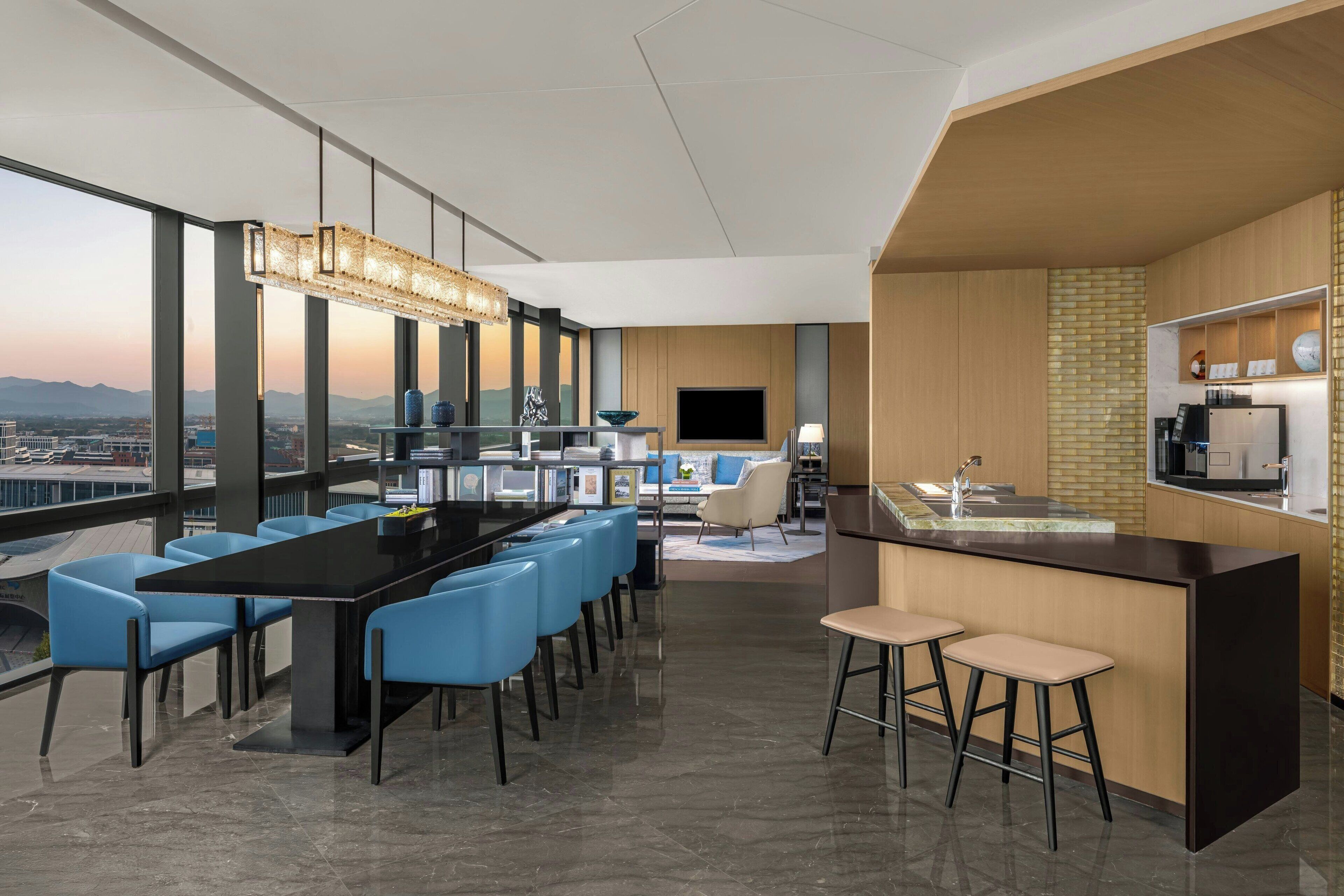 A modern hotel lobby with a long dining table, blue chairs, a bar area, and a large window offering a city view.