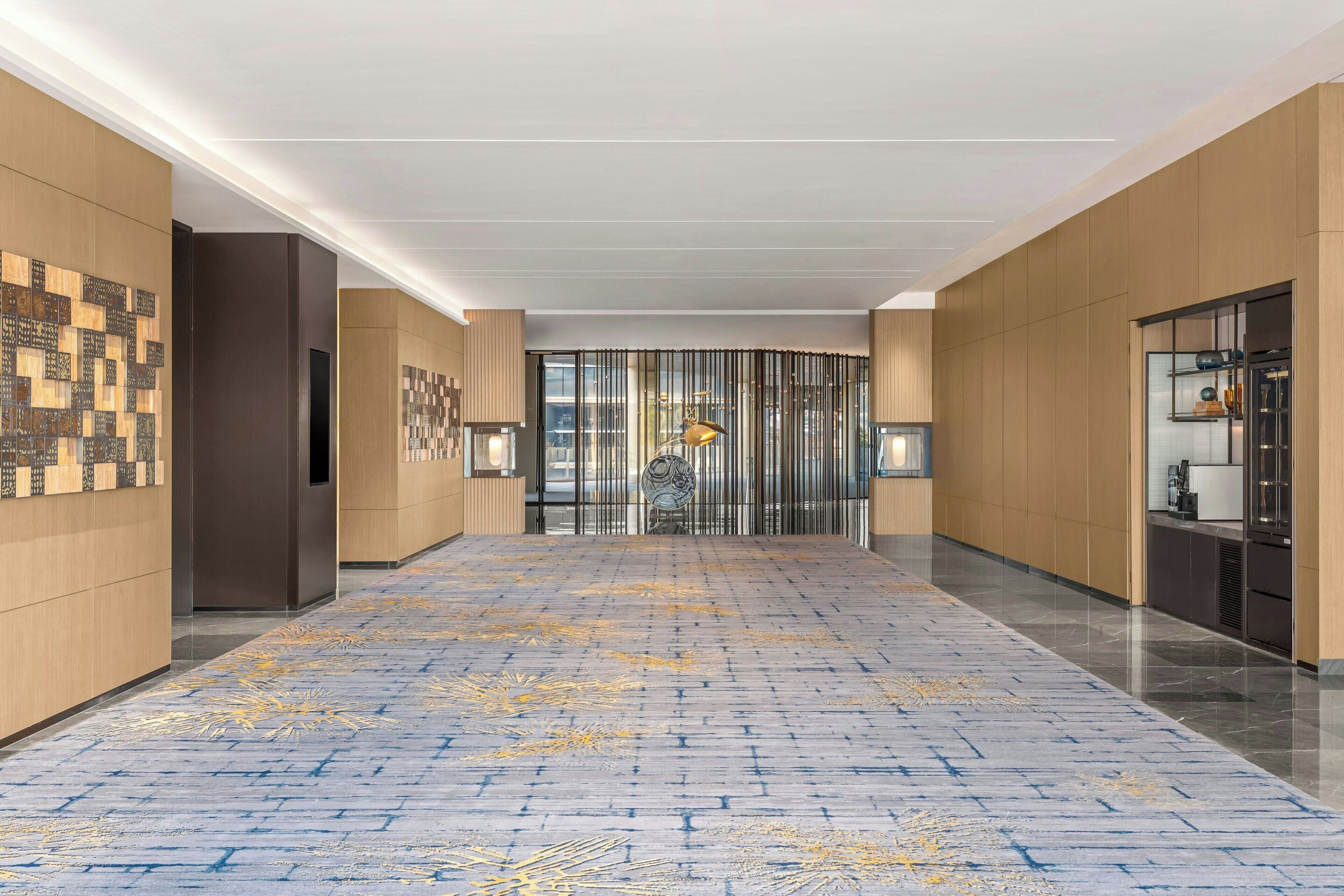 Een moderne hotelgang met een kamerbreed tapijt, houten paneelwanden en een glazen deur aan het einde.