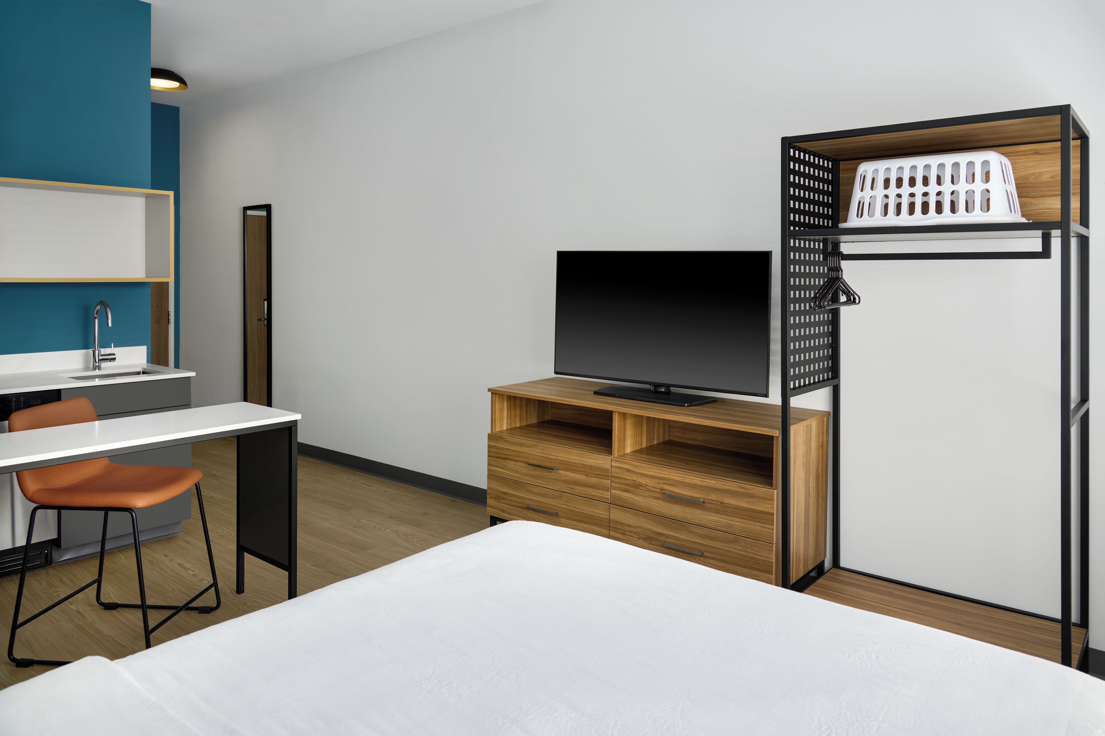 Et moderne hotelværelse med en seng, et skrivebord med stol, et fjernsyn på et træstel og et garderobeskab med en vasketøjskurv.