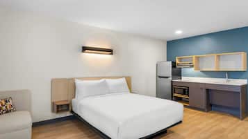 A modern hotel room with a bed, a small sofa, a kitchenette, and a wall-mounted light.