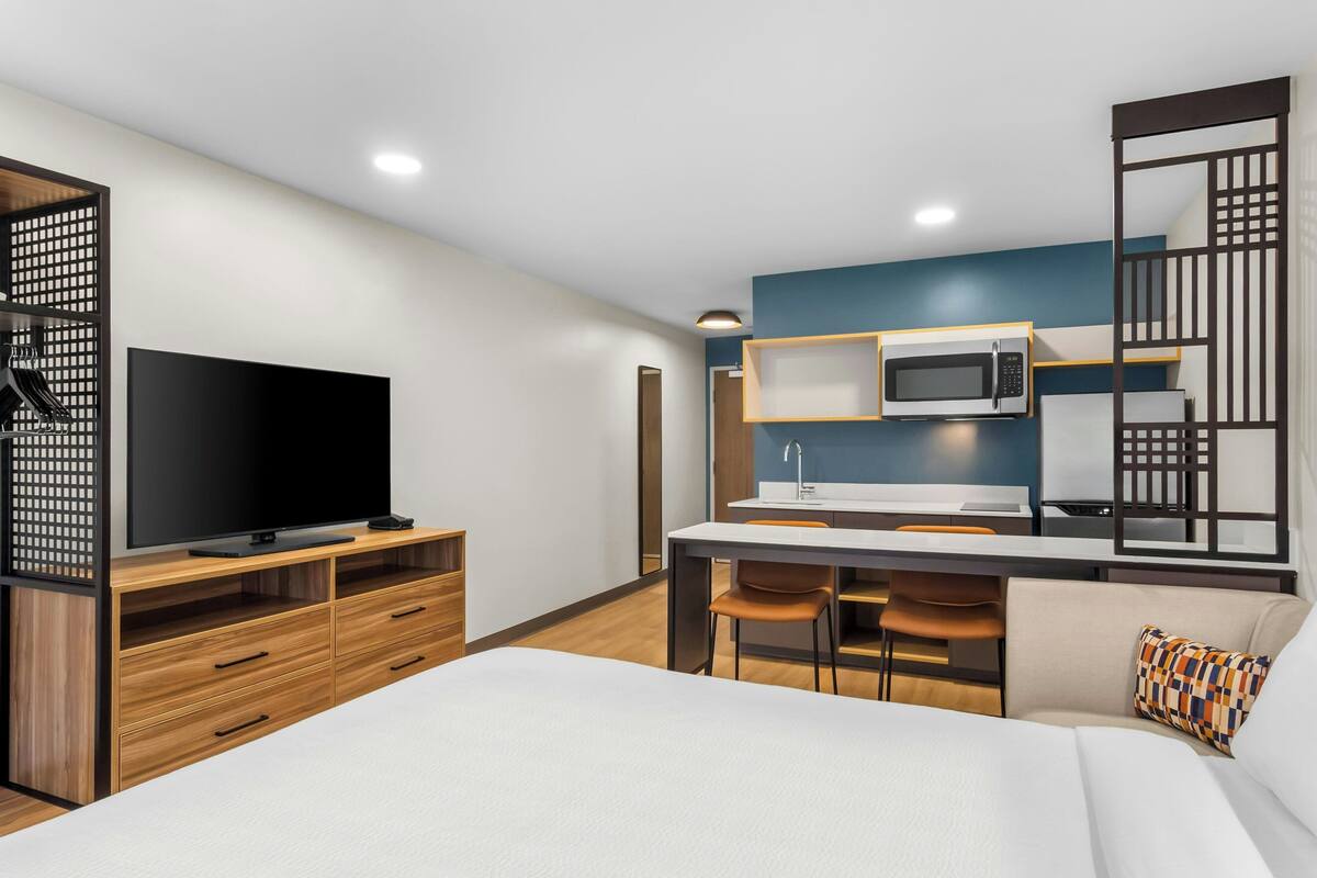 Une chambre d’hôtel moderne dotée d’une télévision à écran plat, de meubles en bois, d’une kitchenette et d’un lit agrémenté d’un oreiller à motifs.