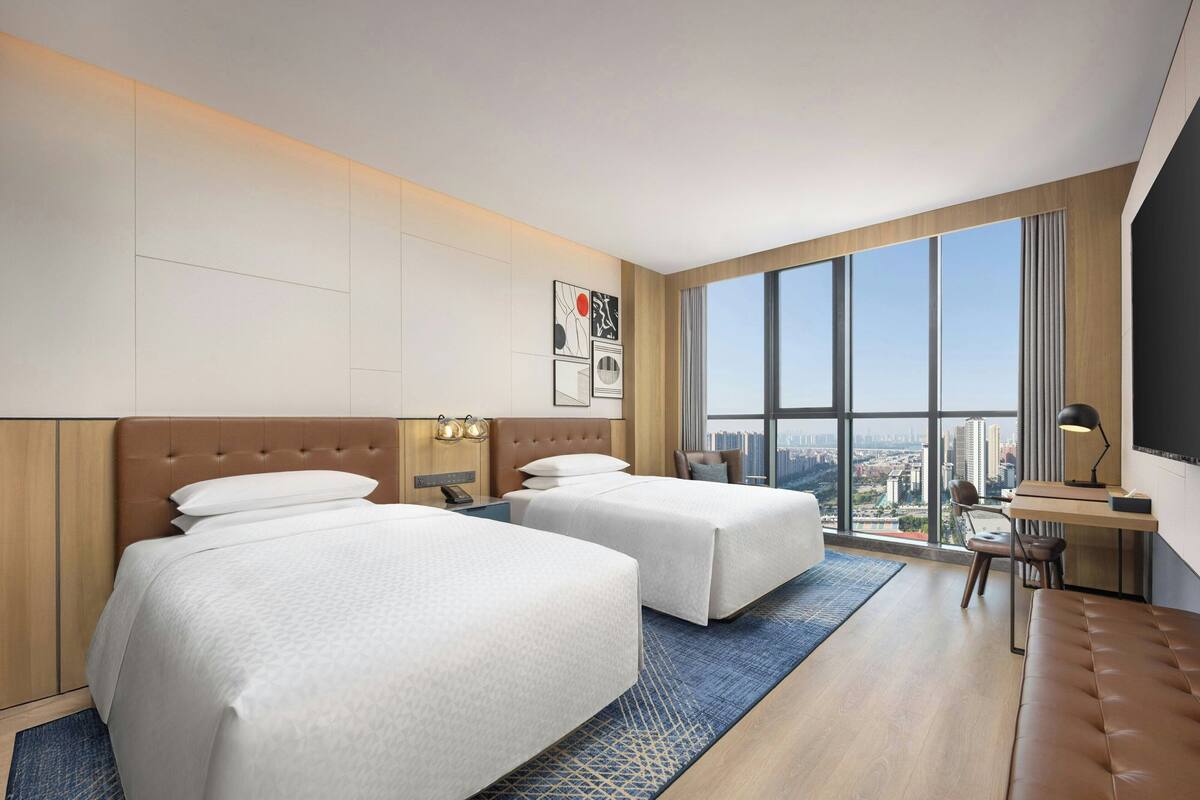 A hotel room with two beds, a desk, a chair, and a large window offering a city view.