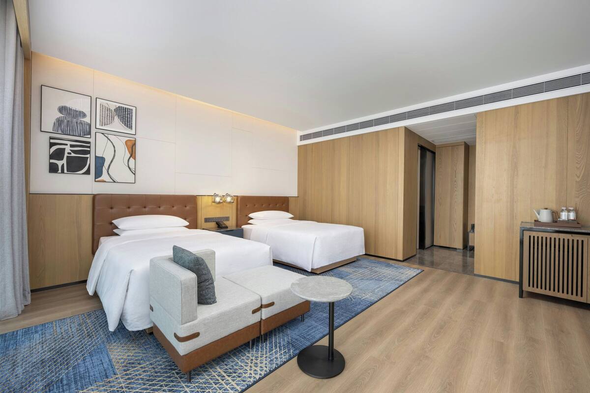 A modern hotel room with two beds, a small table, and artwork on the wall.