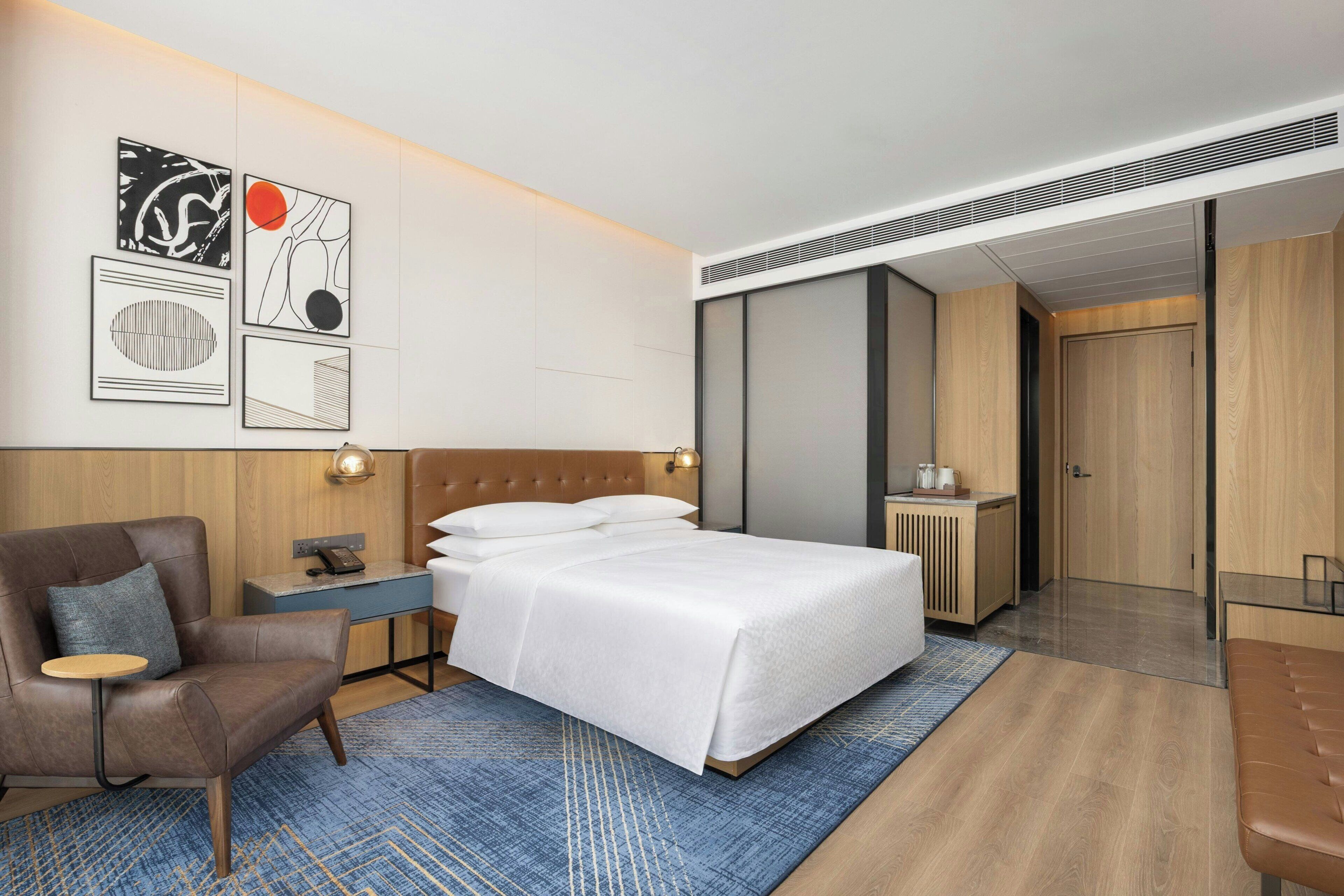 A modern hotel room with a bed, a chair, a desk, and artwork on the wall.