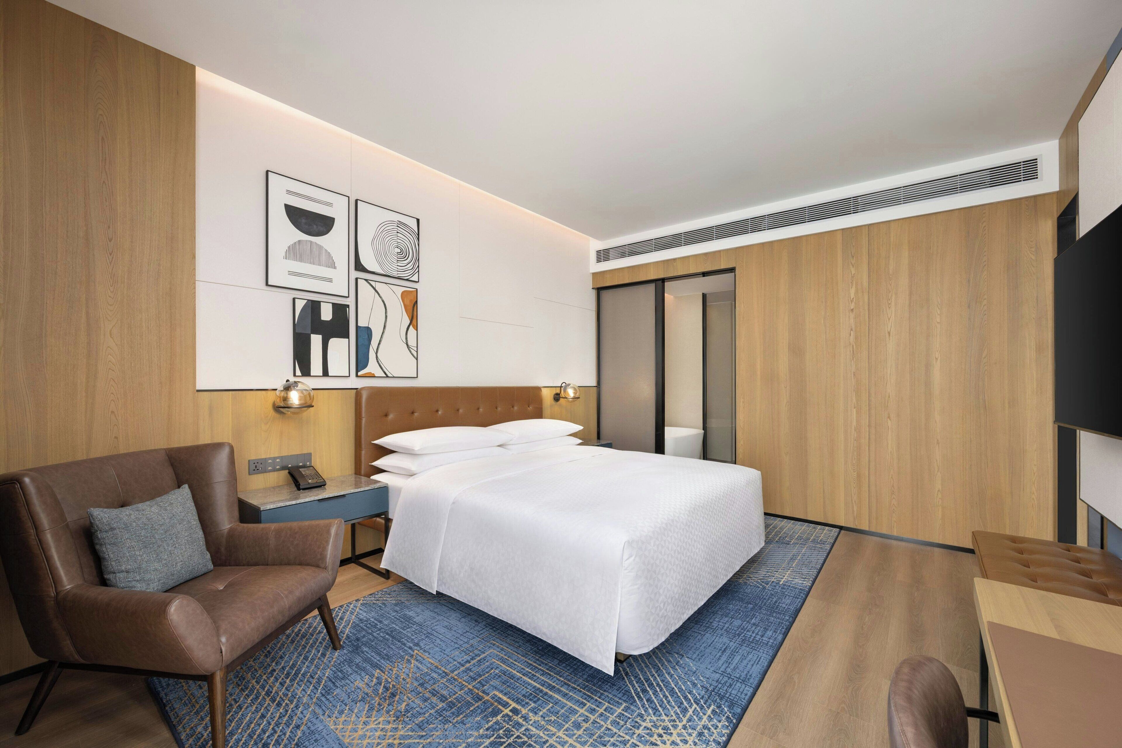 A modern hotel room with a large bed, a leather armchair, a desk, and a television.
