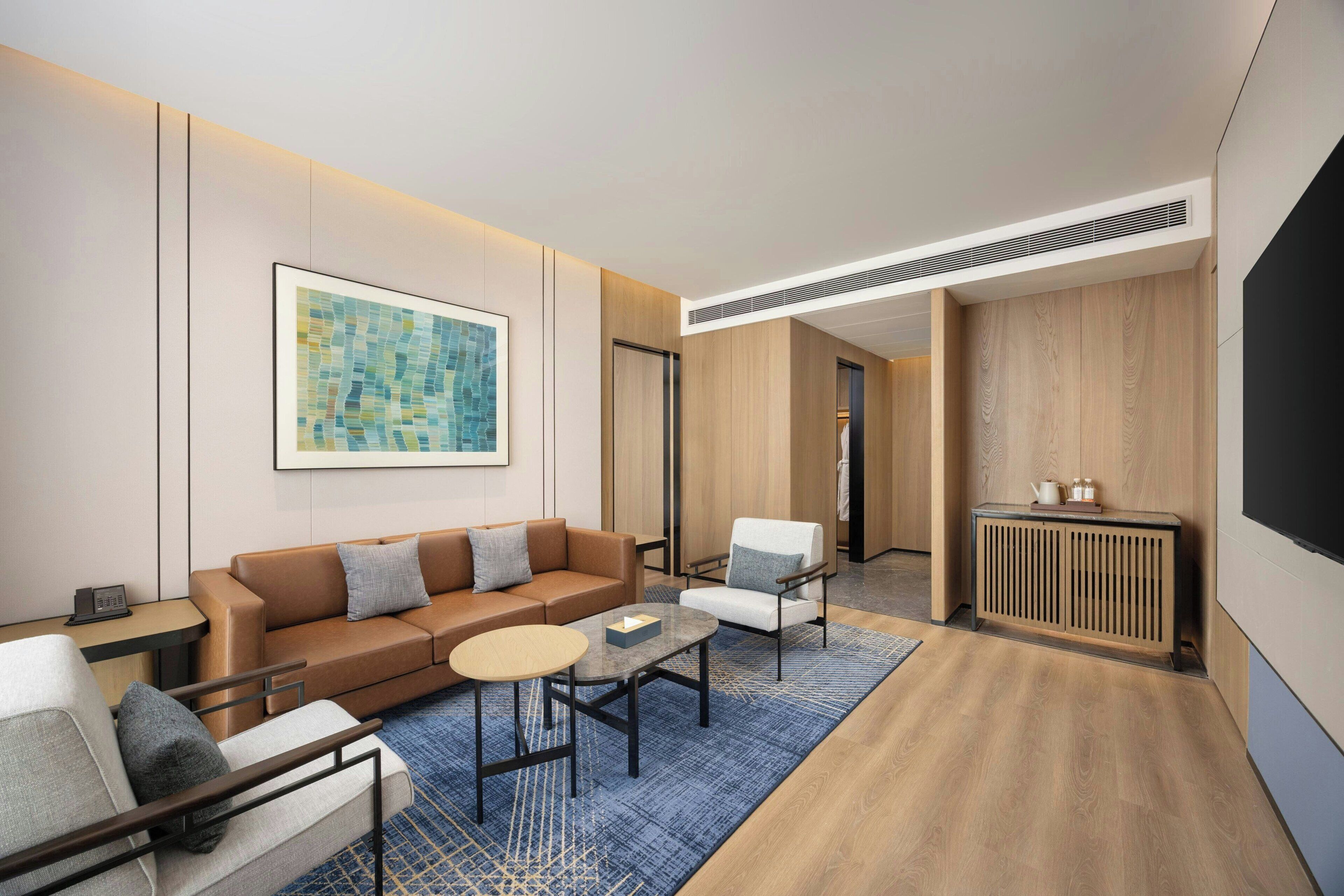 Habitació d'hotel moderna amb un sofà de cuir marró, una taula rodona petita, una butaca blanca i una catifa blava amb dibuixos.