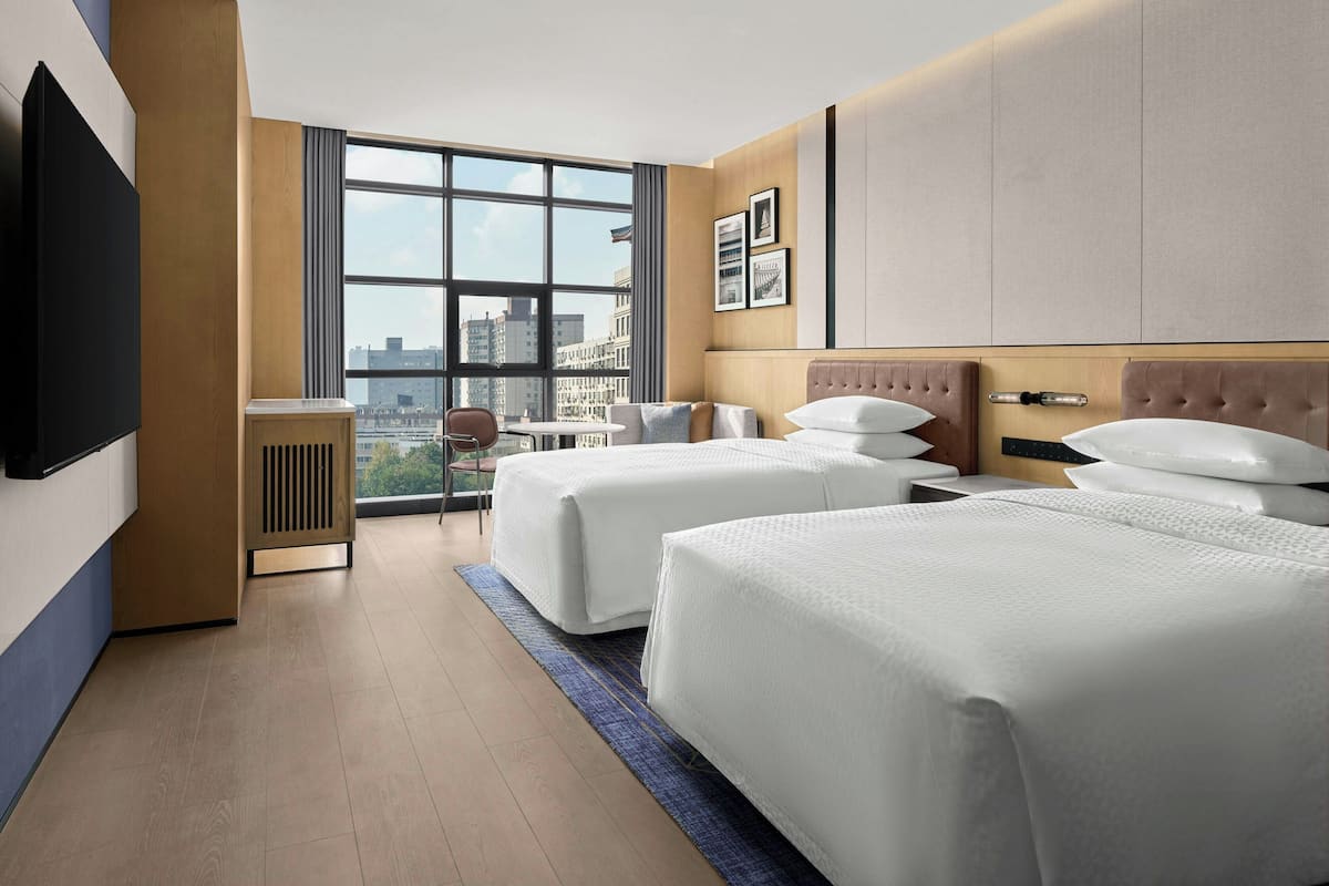 Habitación de hotel con dos camas, televisor, escritorio y ventana grande con vistas a la ciudad.