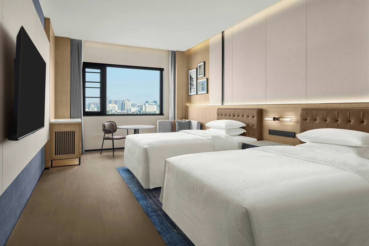 Habitación de hotel con dos camas, un escritorio, una silla, un televisor y vistas a la ciudad.