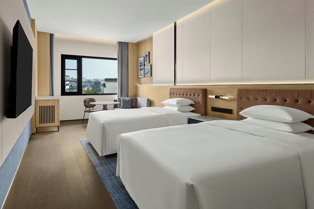 Habitación de hotel con dos camas, televisión, escritorio con silla y ventana con vista a la ciudad.