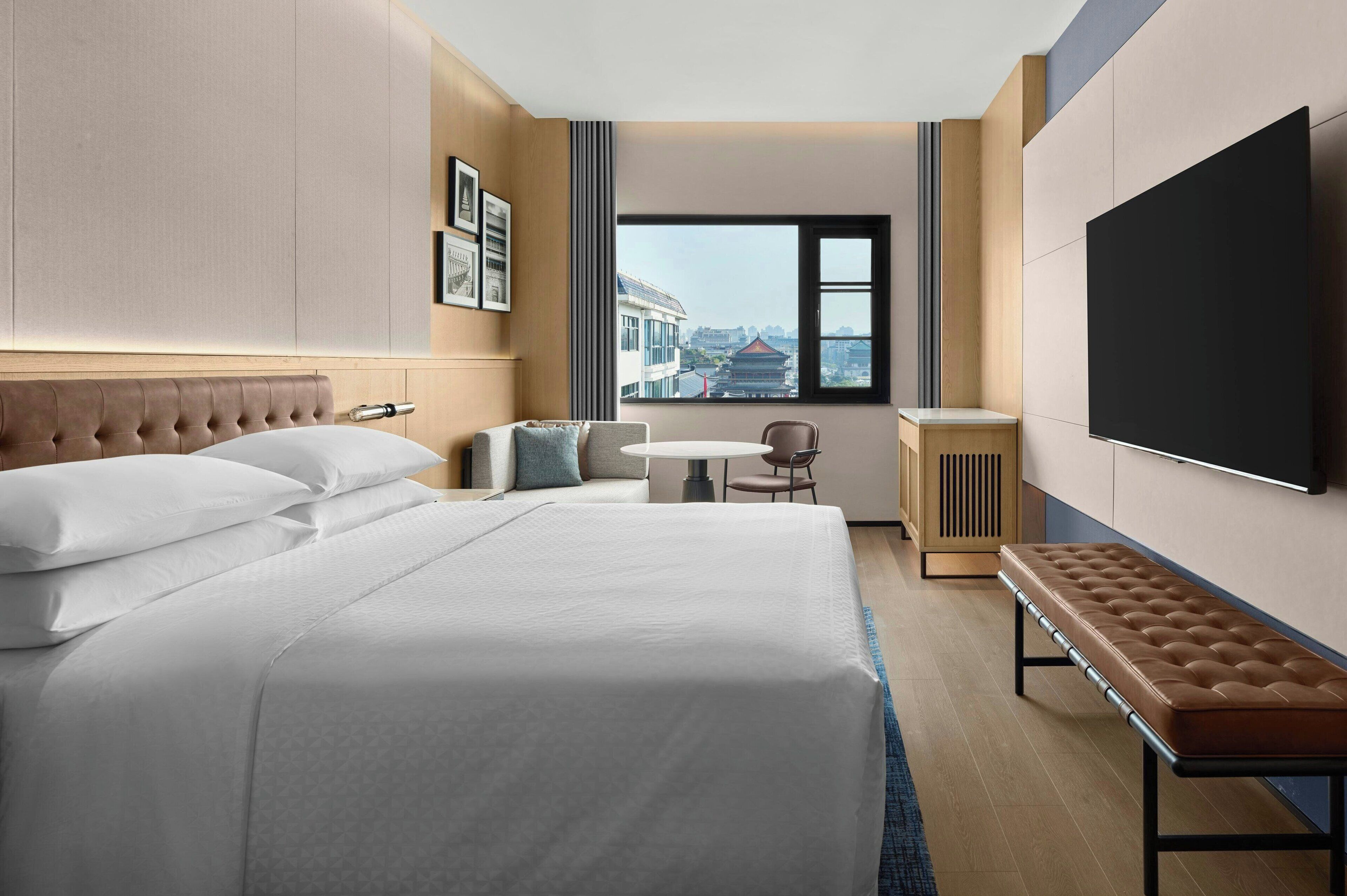 Habitación de hotel con una cama grande, una mesita con una silla, un banco, un televisor y una ventana con vistas a la ciudad.