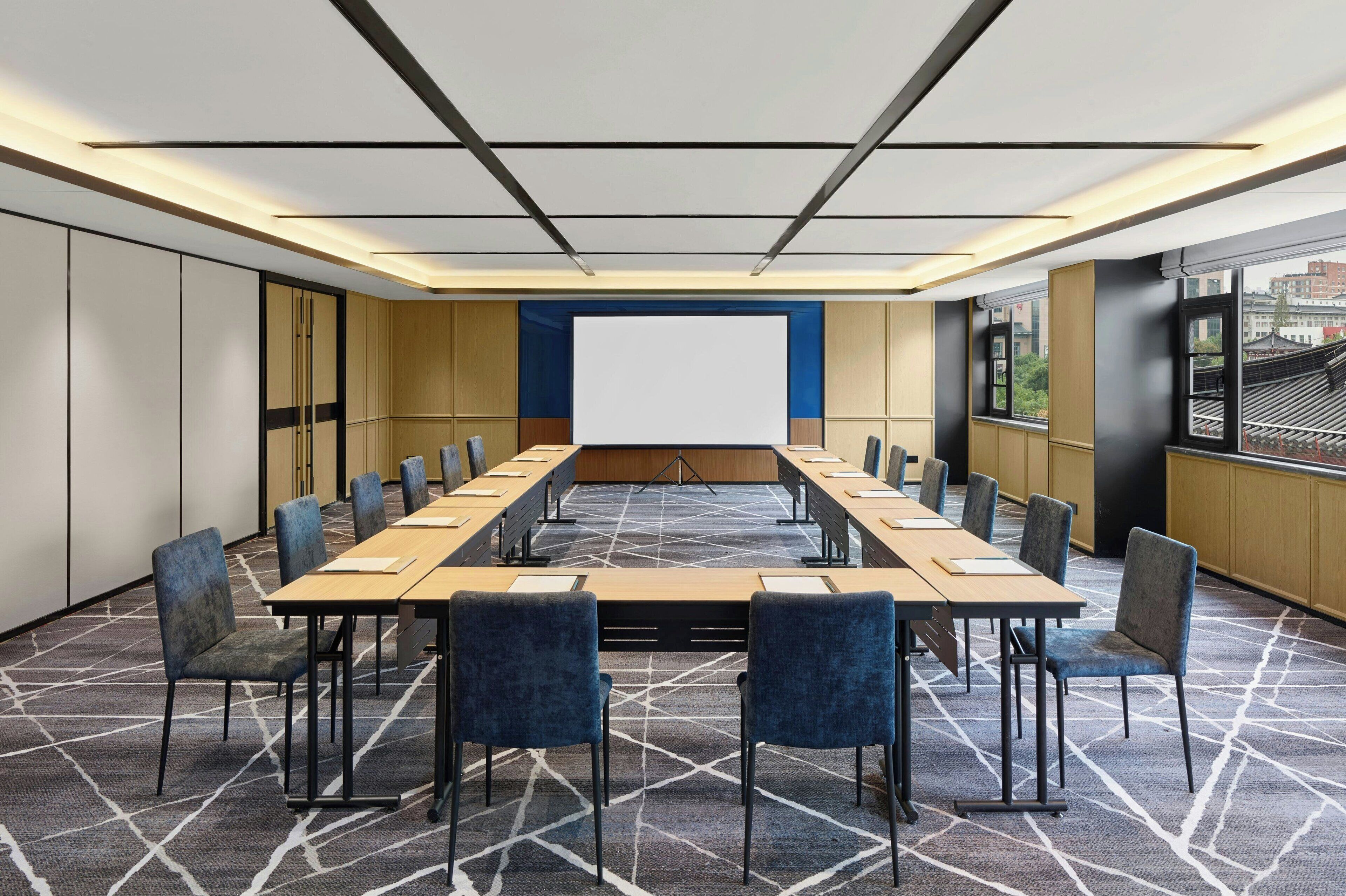 A conference room with a long table, chairs, a projector screen, and a patterned carpet.
