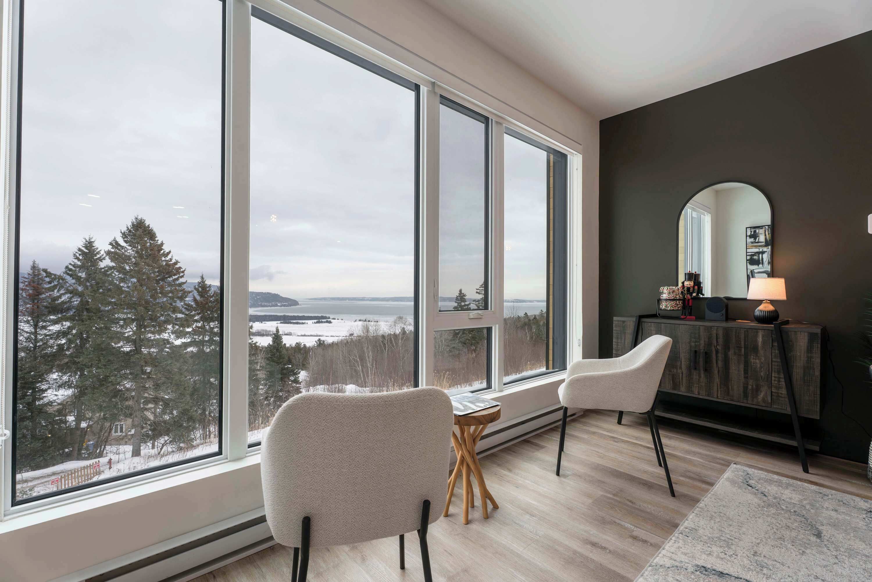 A modern living room with large windows, a fireplace, and a view of a forest and water.