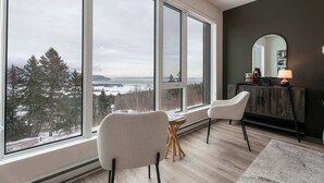 A modern living room with large windows, a fireplace, and a view of a forest and water. - 211 | Bright Condo | Pool and Cozy Atmosphere (Baie-Saint-Paul)