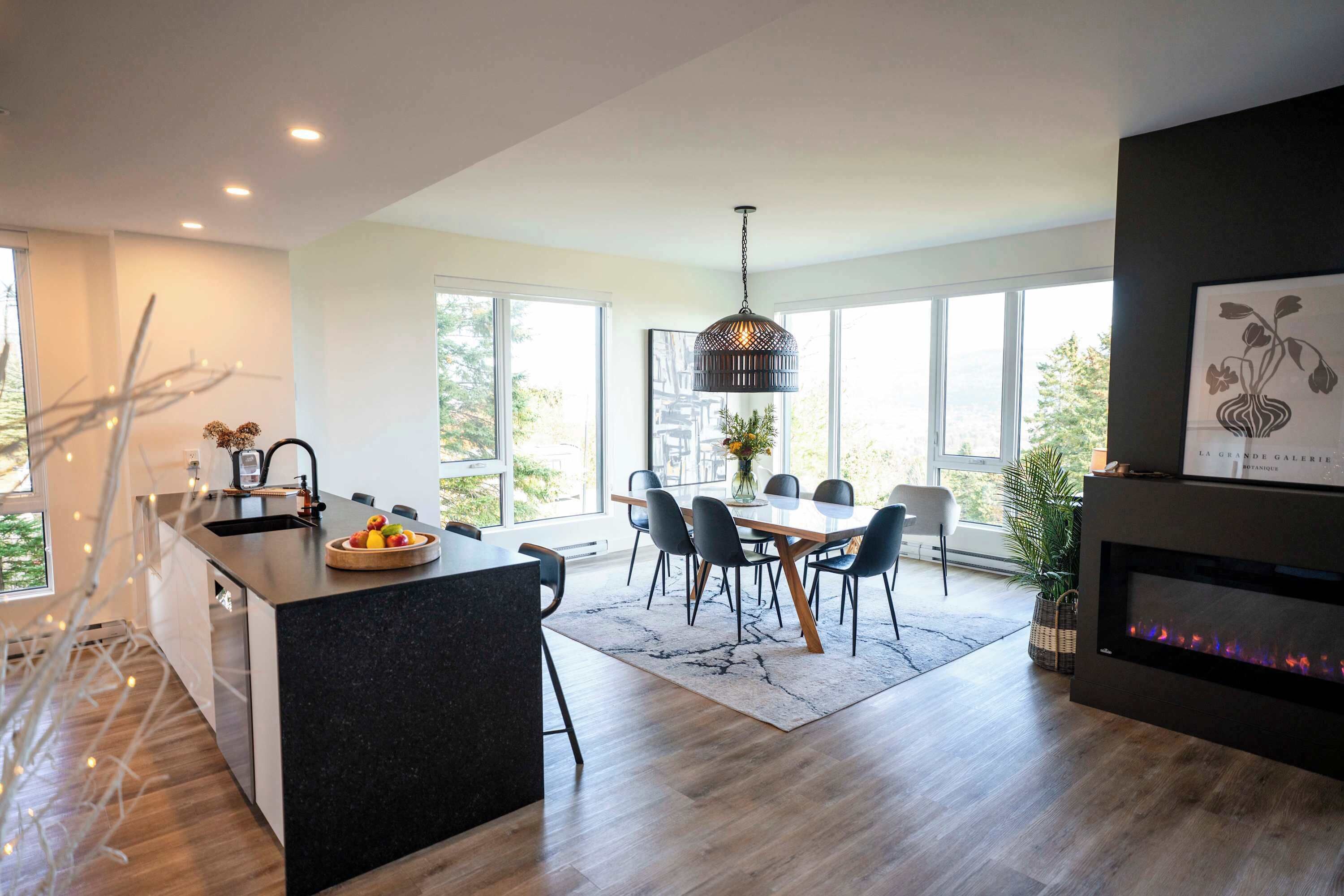 A modern kitchen with a central island, dining area with chairs, large windows, and a fireplace.