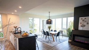 A modern kitchen with a central island, dining area with chairs, large windows, and a fireplace. - 211 | Bright Condo | Pool and Cozy Atmosphere (Baie-Saint-Paul)