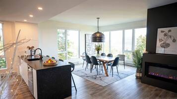 A modern kitchen with a central island, dining area with chairs, large windows, and a fireplace.