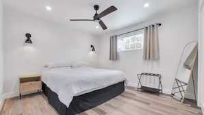 A bedroom with a bed, a ceiling fan, a window with curtains, a bedside table, and a mirror. - Cozy Master King suite- play room- hot tub (Denver)