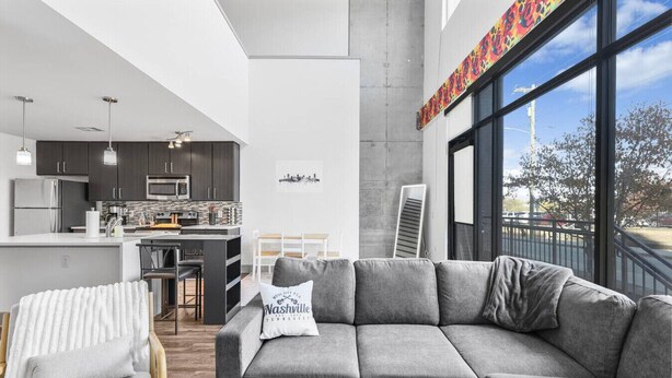 A modern living space with a grey sofa, a kitchen area, and a large window offering a view of the outdoors. - *NEW* Lux Loft V2 (Close to Vandy!) (Nashville)