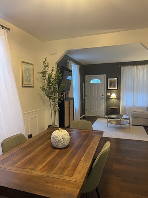 Ein Essbereich mit Holztisch, Stühlen, einer Vase und einer Pflanze. Eine Tür führt in einen anderen Raum mit einer Lampe und einem Sofa.