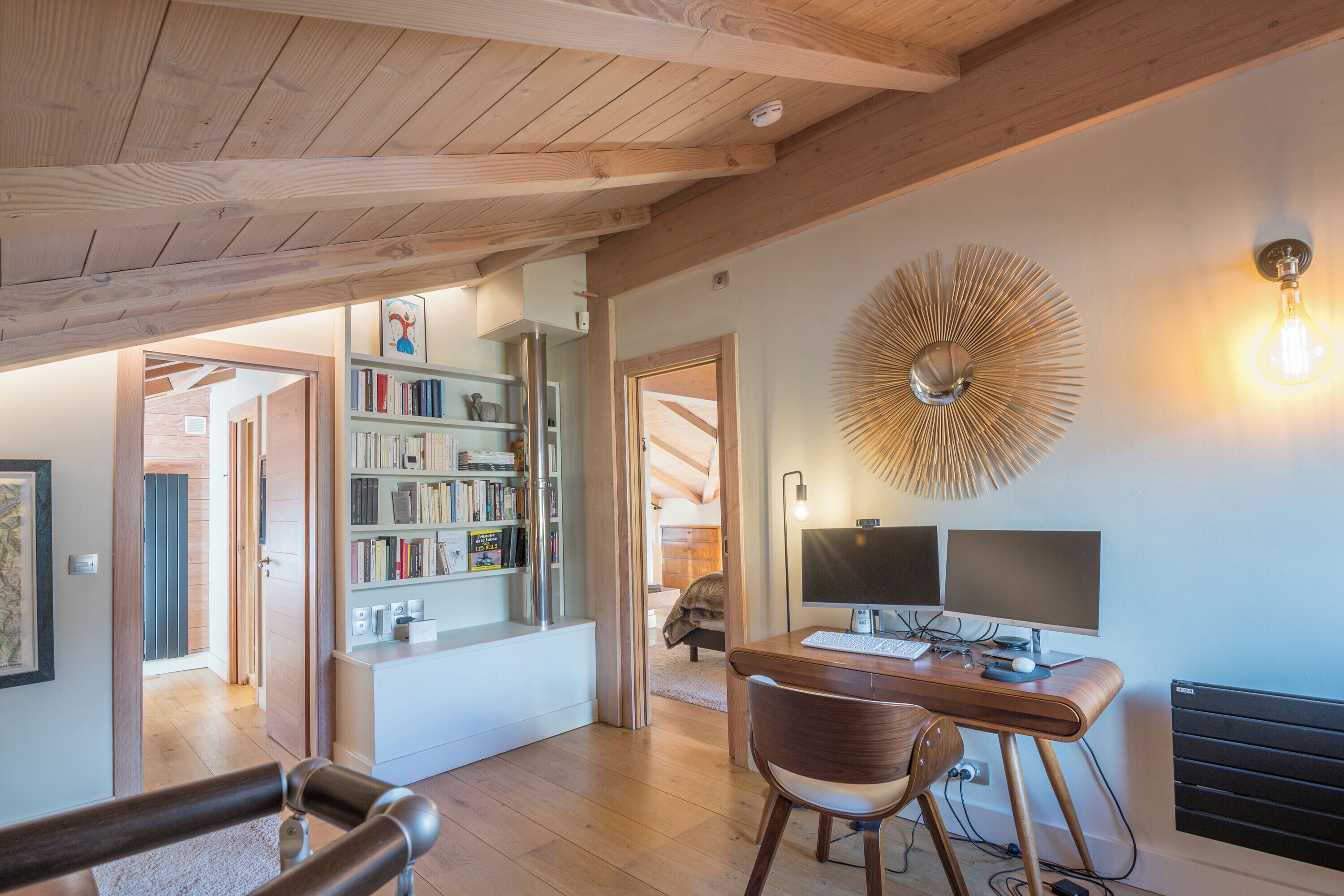 A modern living room with a wooden ceiling, a bookshelf, a desk with two monitors, and a decorative light fixture.