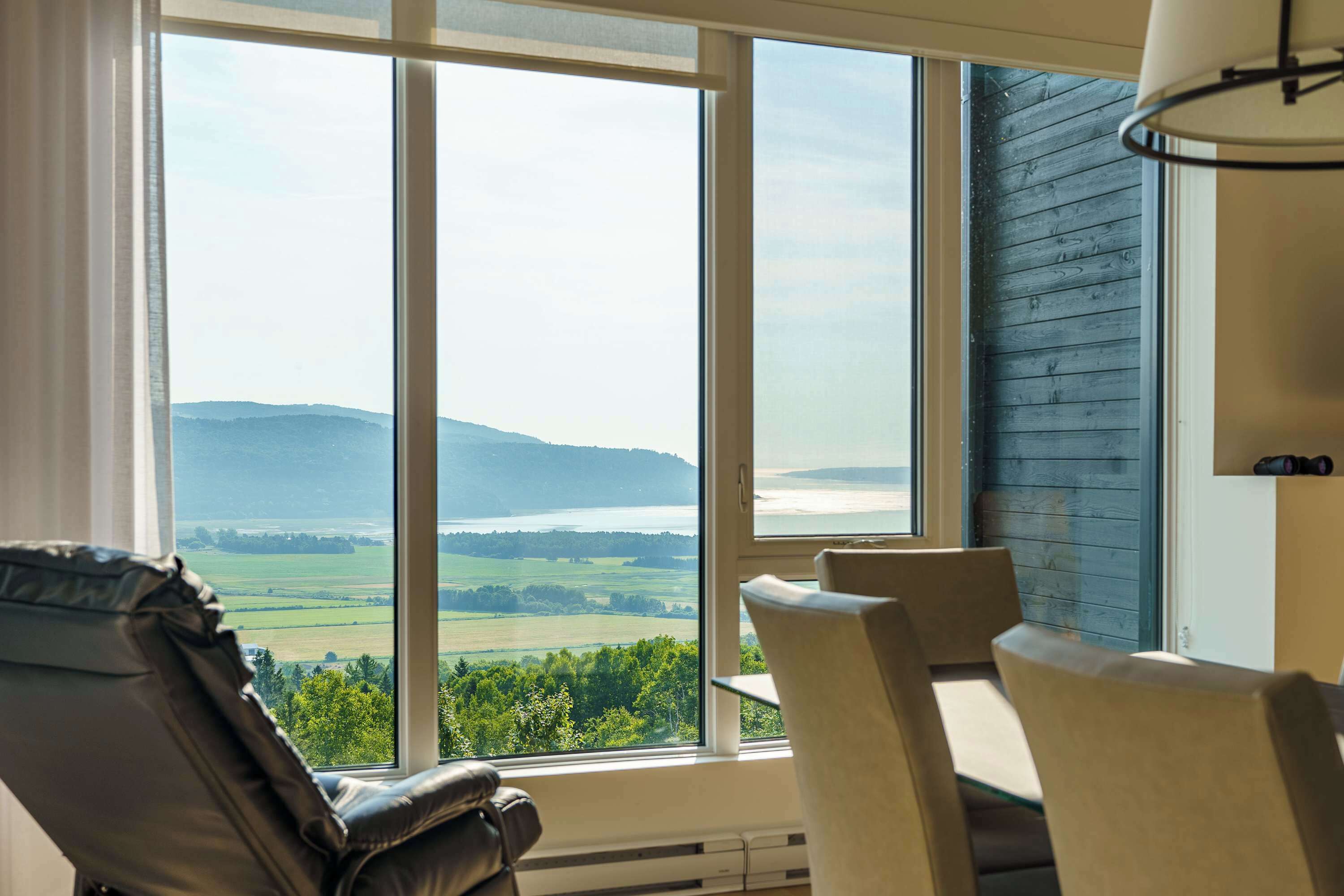 A room with a large window offering a view of a landscape with hills and a body of water. There are two chairs positioned near the window.