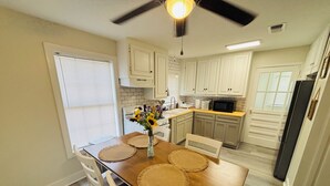Cozinha com armários brancos, uma mesa de jantar de madeira com jogos americanos, micro-ondas e ventilador de teto.
