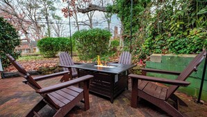 Un patio con una mesa y cuatro sillas tipo Adirondack, rodeado de follaje y un suelo de ladrillo.