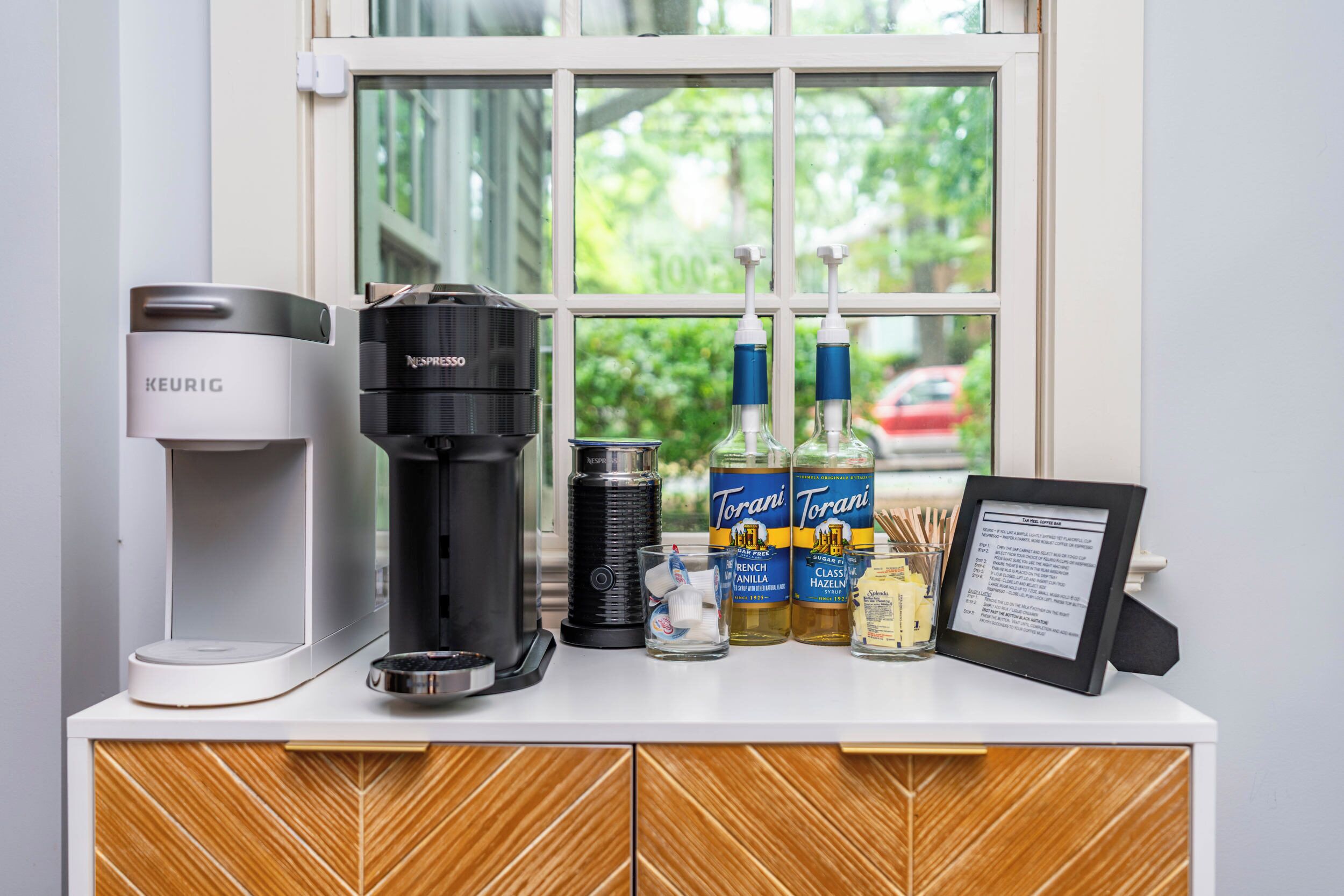 Una estación de café con una máquina Keurig, una máquina Nespresso y siropes Torani.