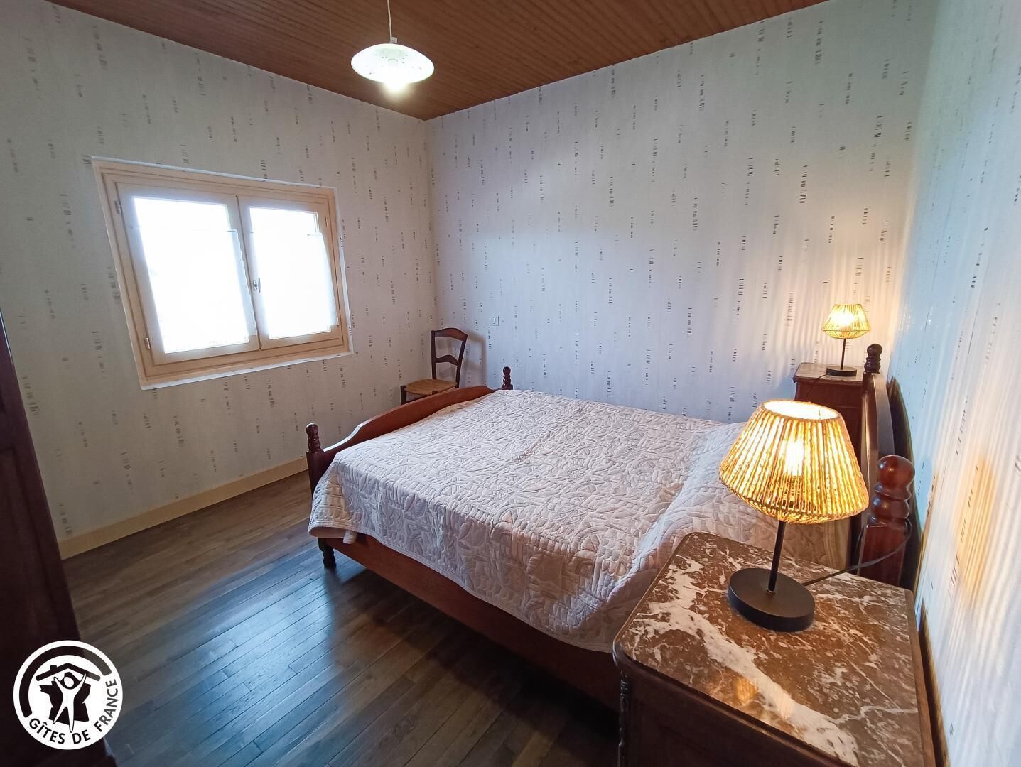 A bedroom with a wooden bed, a nightstand with a lamp, a chair, and a window with blinds.