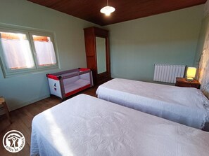 Ein Zimmer mit zwei Betten, einem Babybett, einem Fenster mit Vorhängen, einem Heizkörper und einer Lampe auf einem Nachttisch.