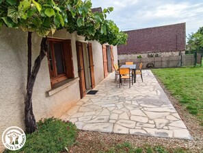 Eine Steinterrasse mit Tisch und Stühlen, ein kleines Fenster und ein Baum mit grünen Blättern.