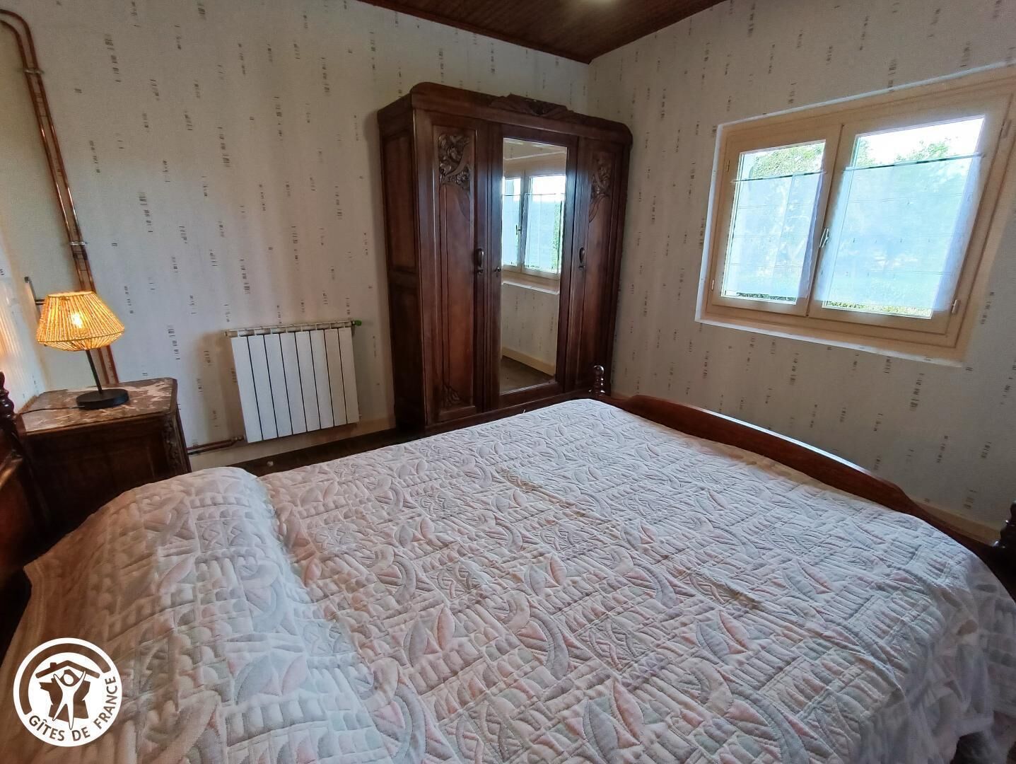 A bedroom with a large bed, a wooden wardrobe, a radiator, and a window with a view of greenery.