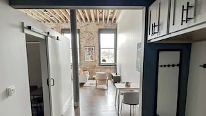 A modern interior with a wooden floor, white walls, and a brick accent wall. There is a dining area with a table and chairs, and a large window providing natural light. - Sleepover | Modern 1BD/1BA Loft - Newton (Newton)