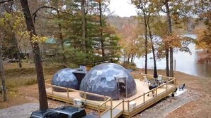 Une structure en forme de dôme surmontée d’une terrasse en bois, un véhicule garé à proximité et un lac en arrière-plan.