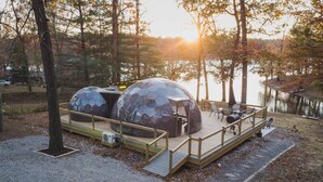 Une cabane dotée d’une terrasse et de deux structures sphériques, probablement destinées à la détente ou aux repas, située près d’une étendue d’eau.