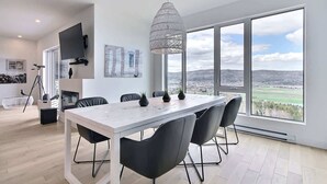 A modern dining area with a white table and black chairs, large windows with a view of the countryside, and a hanging light fixture.