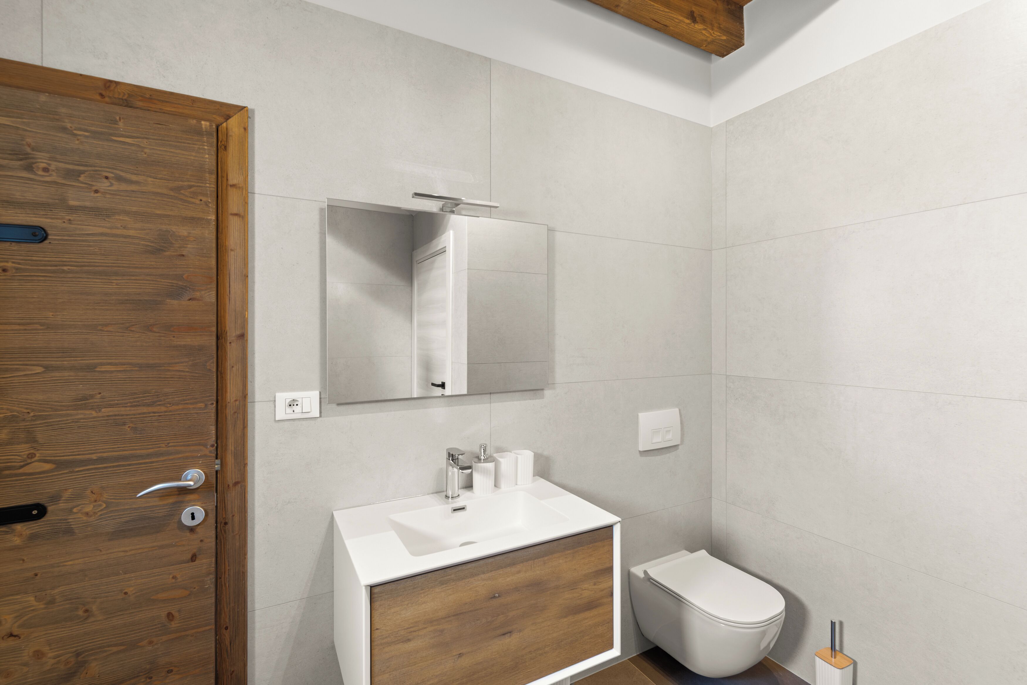 Un bagno moderno con una porta in legno, un lavabo bianco e uno specchio sopra, e un water.