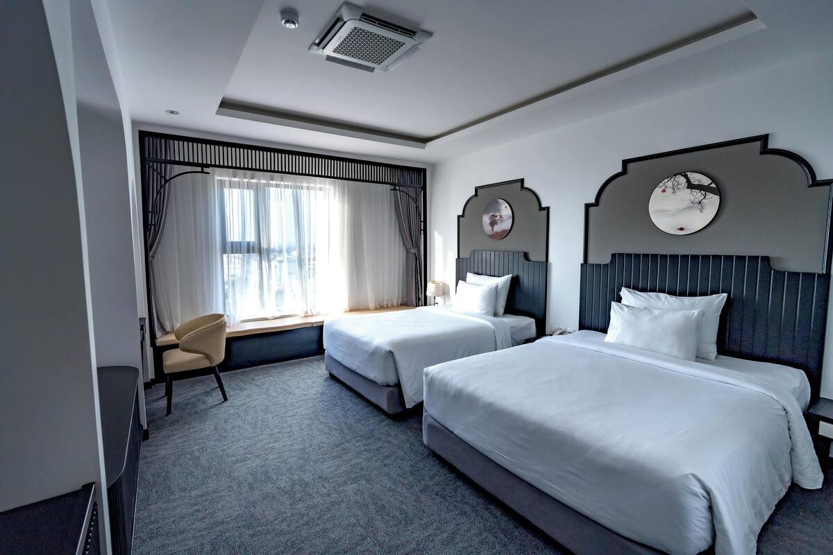 Habitación de hotel con dos camas, una silla, un ventanal con cortinas y un reloj de pared.