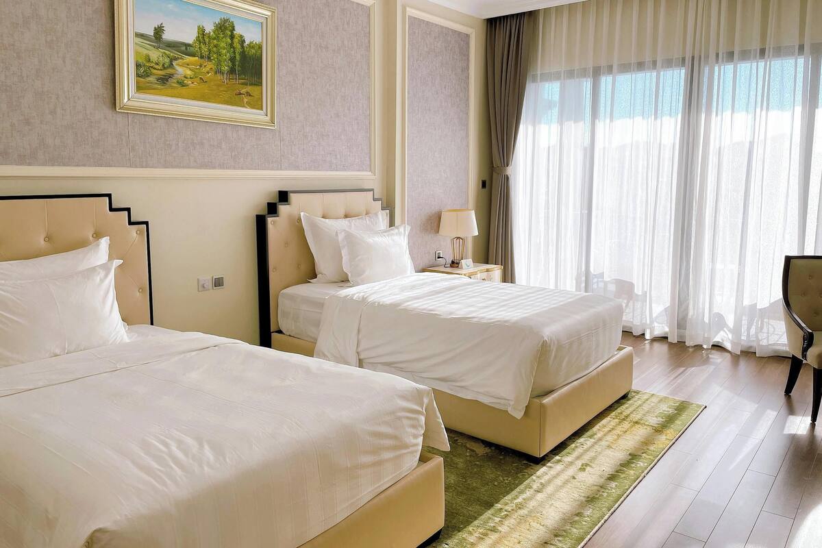 Habitación de hotel con dos camas, un cuadro en la pared, una ventana con cortinas y una alfombra verde.
