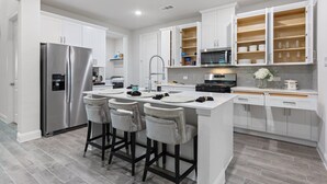 A modern kitchen with white cabinets, a stainless steel refrigerator, a central island with bar stools, and a microwave above the island. - Where Family Adventures Begin (Seagoville)