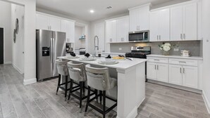 A modern kitchen with white cabinetry, stainless steel appliances, and a central island with bar stools. - Where Family Adventures Begin (Seagoville)