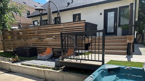 A backyard with a hot tub, wooden deck, outdoor seating, and a house in the background.