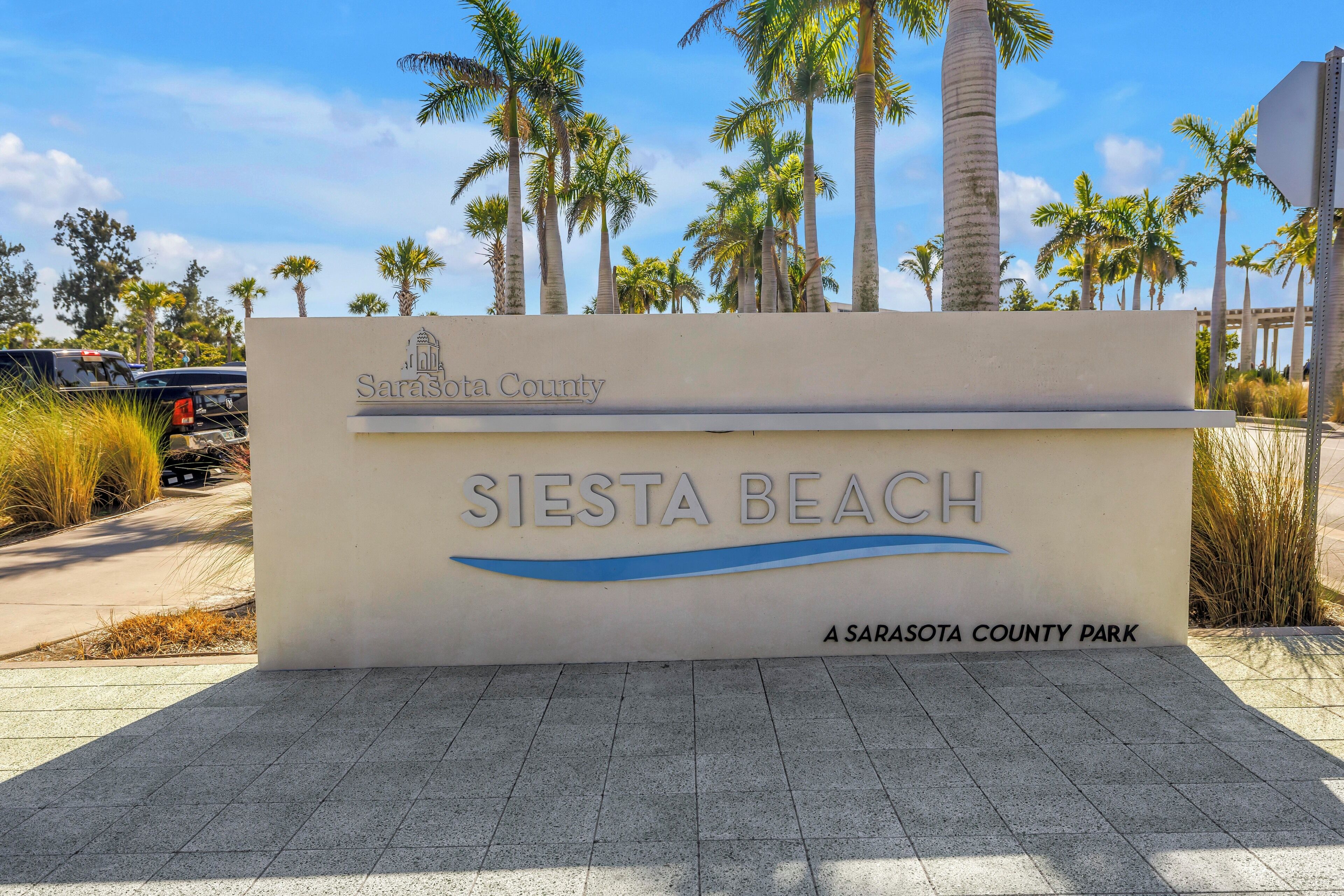 A sign for Siesta Beach, a Sarasota County park, with palm trees and clear skies in the background.