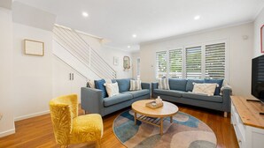 Een moderne woonkamer met een blauwe zithoek, een gele fauteuil, een houten salontafel en een tv op een houten dressoir.