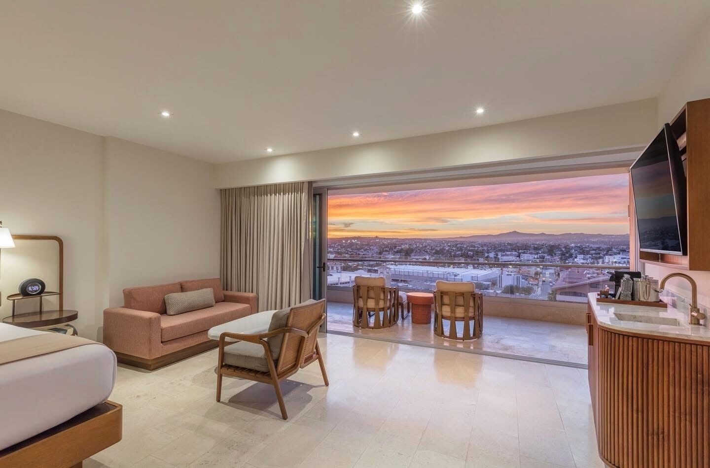 A hotel room with a large window offering a city view, a sofa, a dining table with chairs, a TV, and a bathroom area.