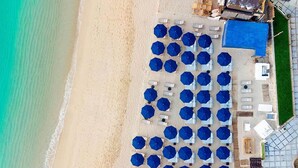 An aerial view of a beachside hotel with rows of blue umbrellas and white lounge chairs, a pool area, and a building with a blue roof. - Corazon Cabo, Infinty Two Queen Sunset View Hot Tub, Spend New Year's in Cabo (Cabo San Lucas)