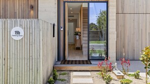 Een modern huis met een houten gevel, een glazen deuropening en een kleine tuin met planten en een bankje.