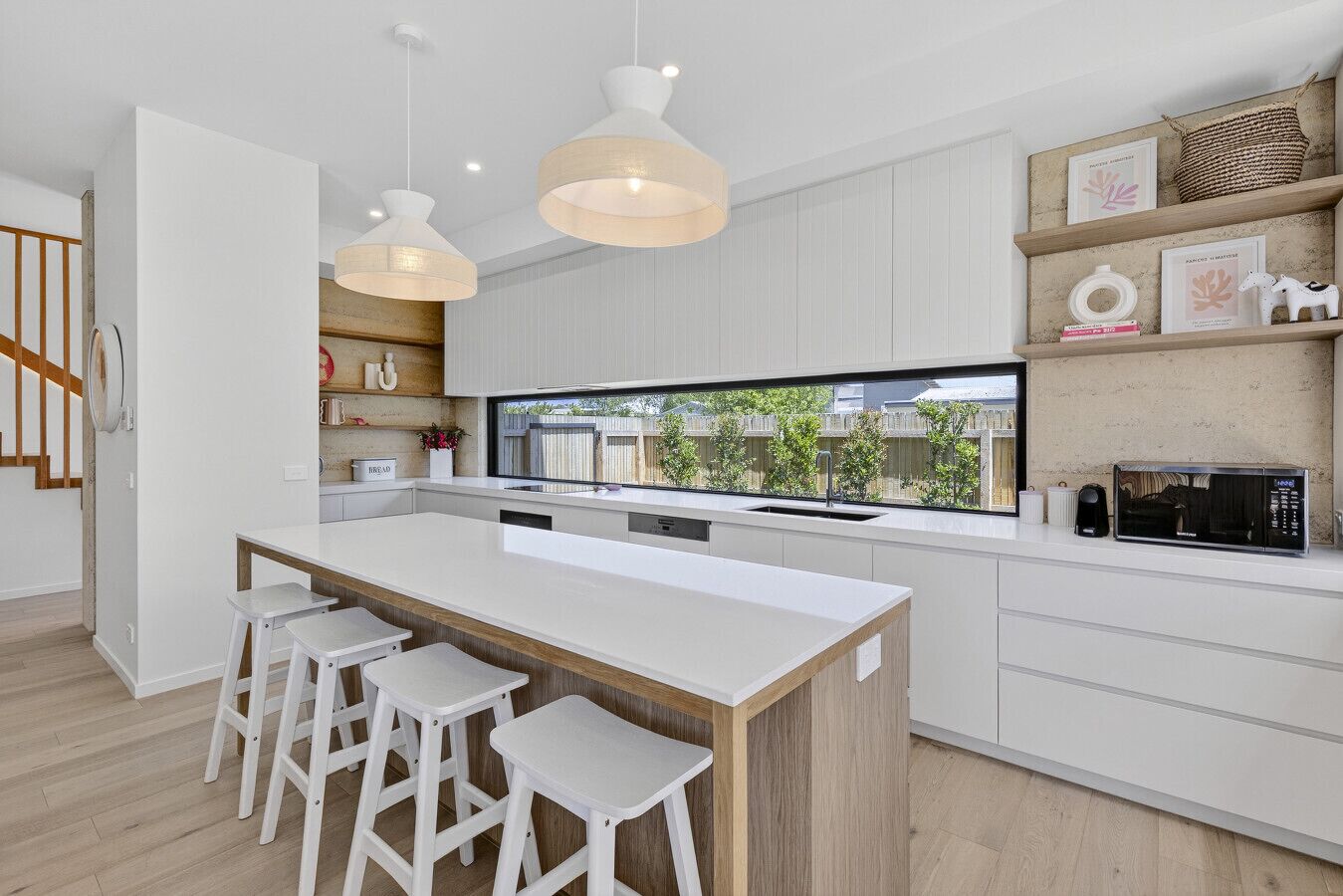 A modern kitchen with a central island, white bar stools, pendant lights, and a view of the outside.
