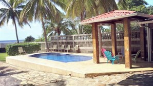 A swimming pool with a wooden deck, lounge chairs, and a covered seating area with a view of the ocean.